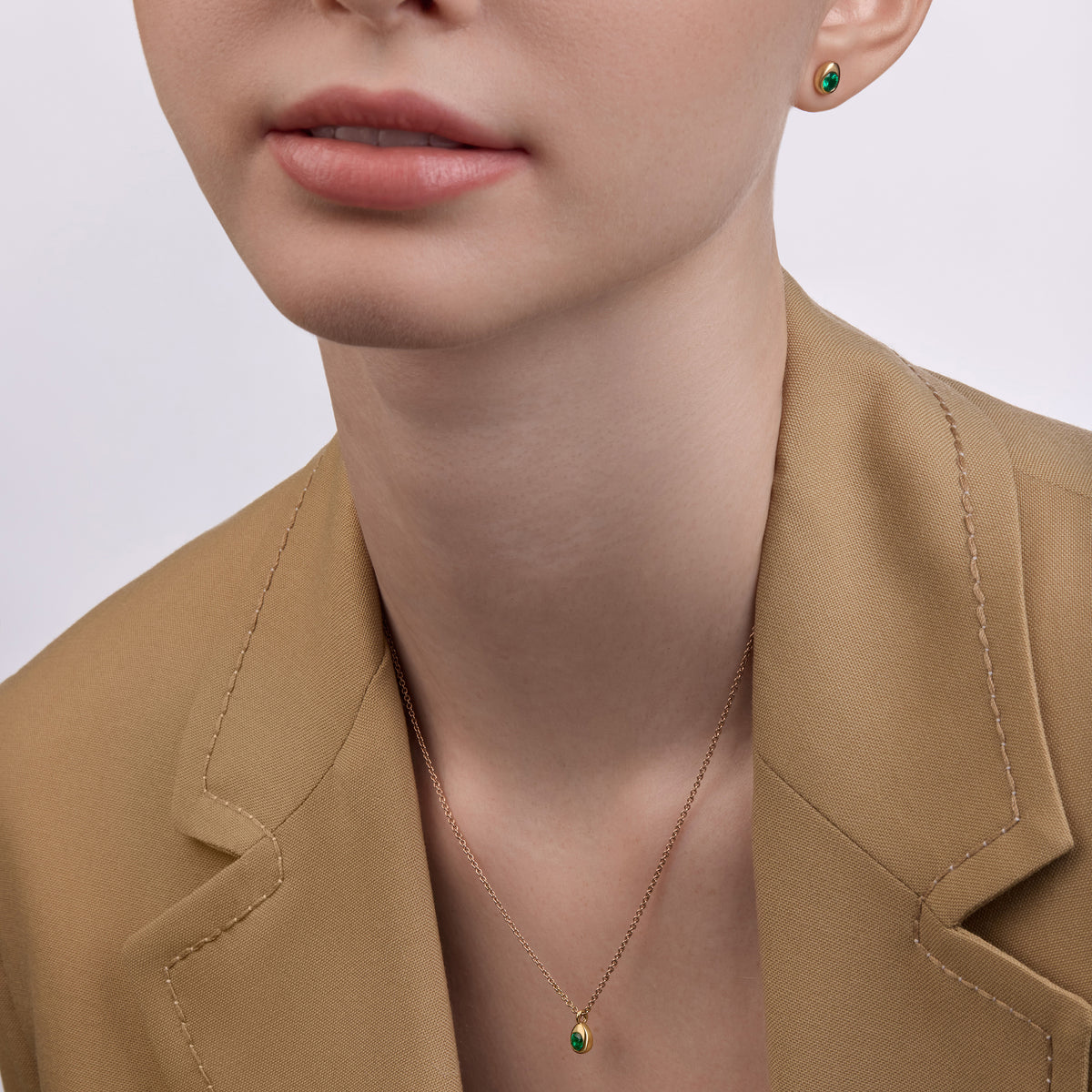 Model wearing Fabergé Code yellow gold emerald stud earrings and matching pendant necklace with a beige blazer.