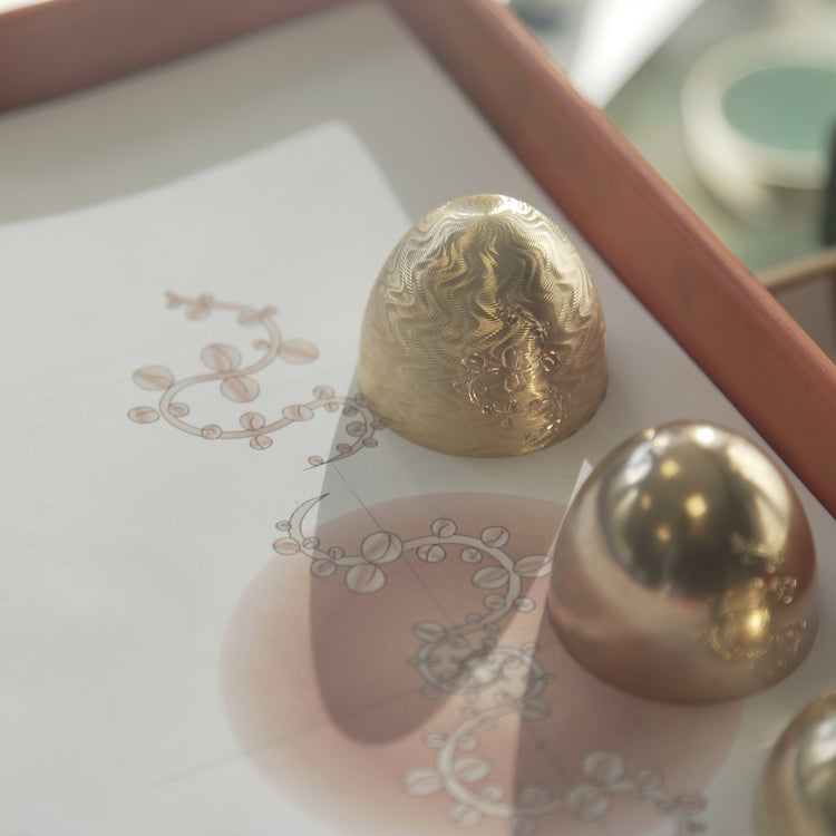 Close-up of Fabergé enamel gold egg-shaped jewellery forms displayed on top of hand-drawn design sketches, highlighting the craftsmanship and heritage-inspired design process.