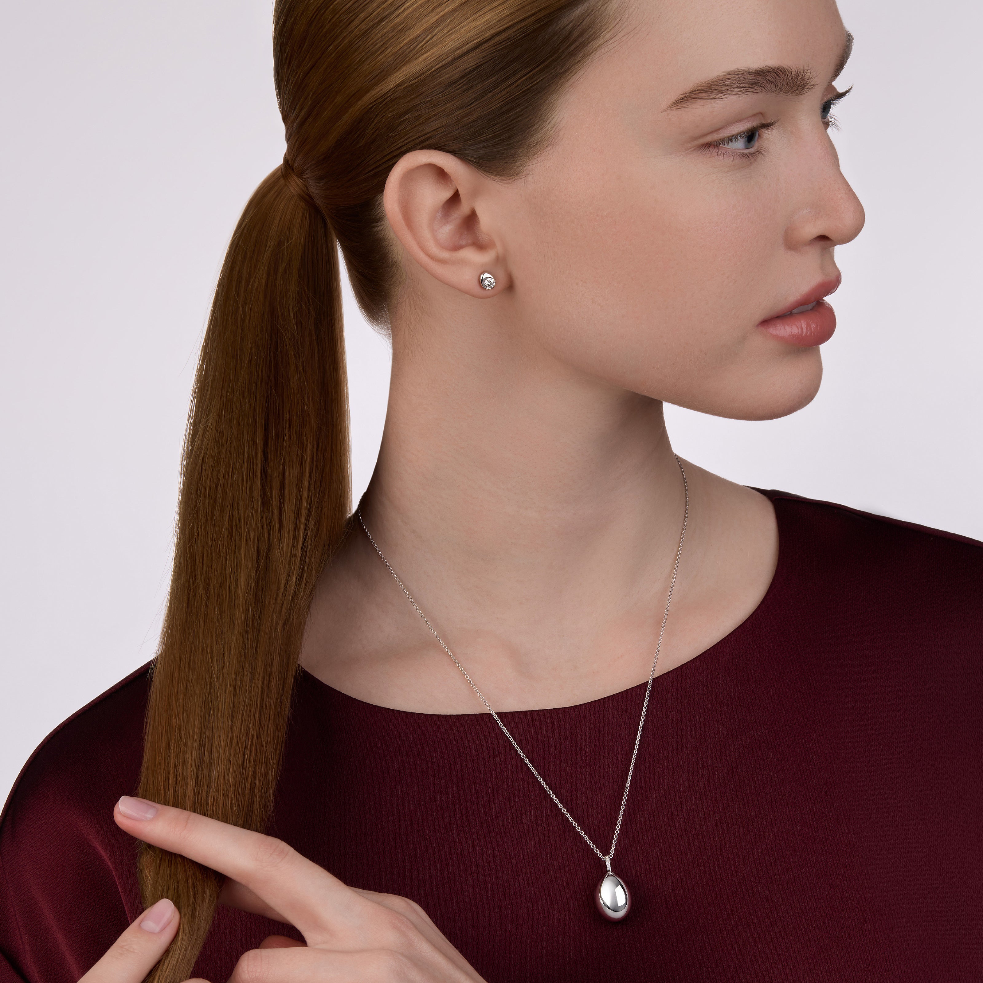 Side profile of a model wearing the Fabergé Essence Petite Pendant necklace and Code diamond stud earrings, styled with a deep burgundy dress against a soft neutral background.