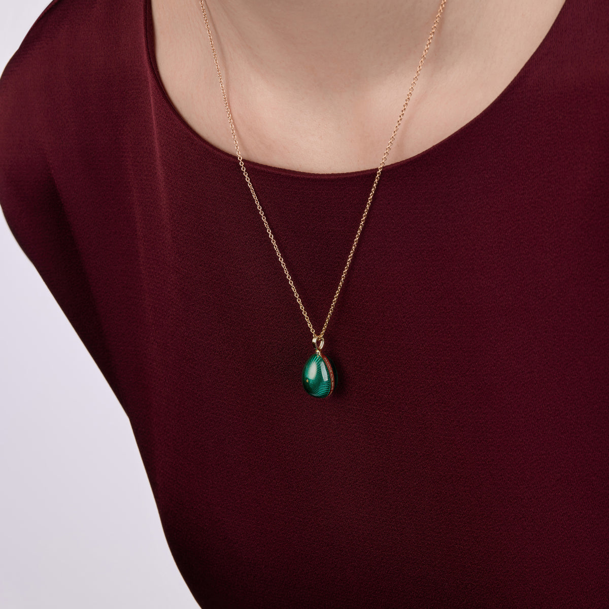 Close-up of a model wearing a yellow gold Fabergé Forever Starburst egg pendant with green enamel detailing, styled against a burgundy satin dress.