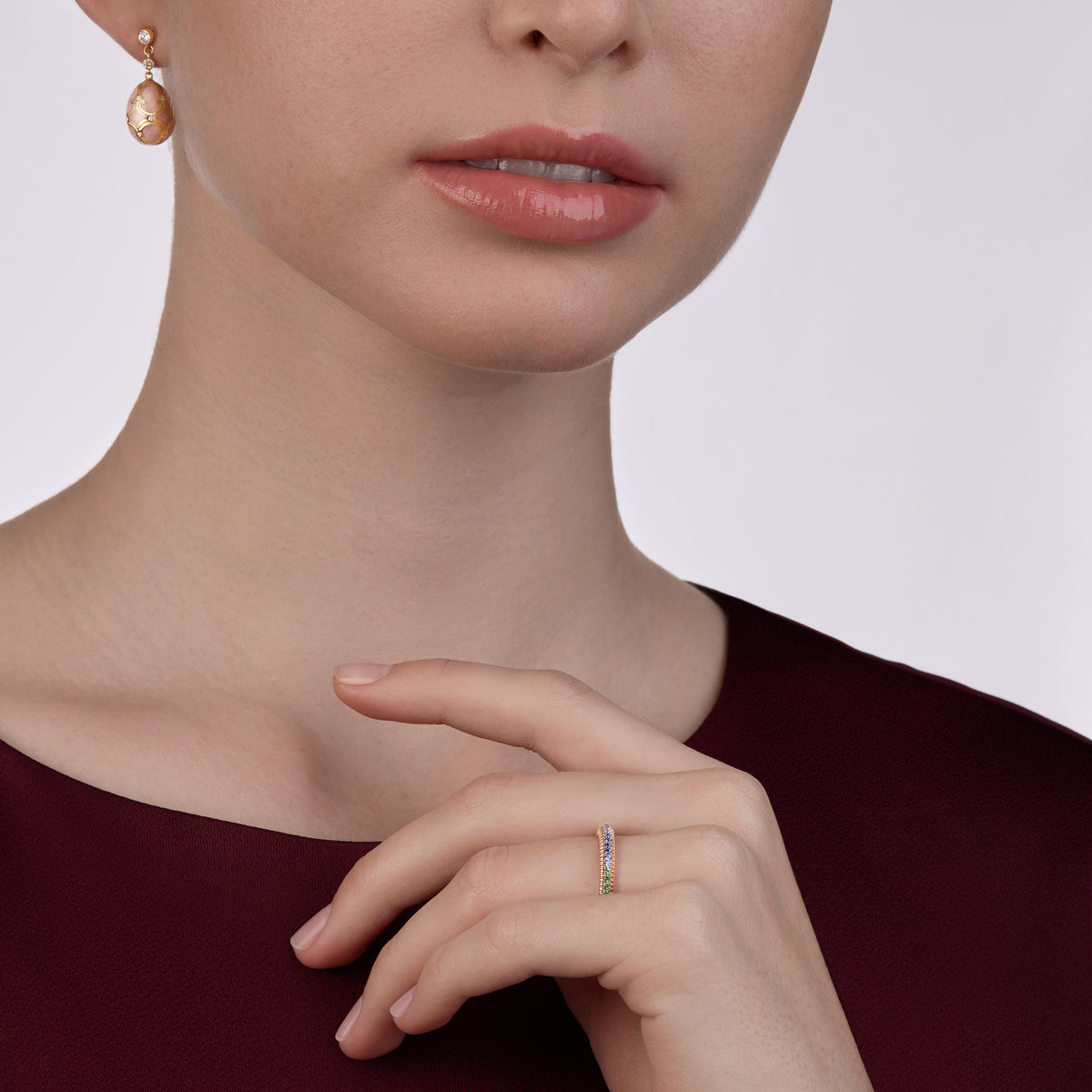 Model wearing Fabergé Heritage egg drop earrings and a yellow gold Love ring set with mixed gemstones, styled against a burgundy satin dress.
