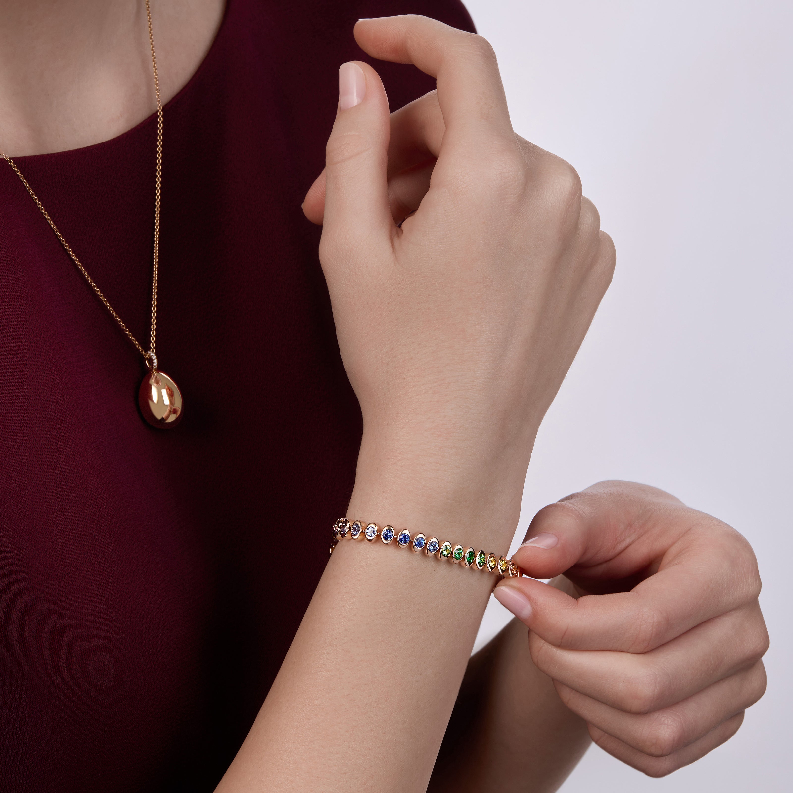 Close-up of a model wearing the Fabergé Heritage Forever Mini Egg Pendant necklace and a Fabergé Code tennis bracelet with multicoloured gemstones, styled with a deep burgundy dress.