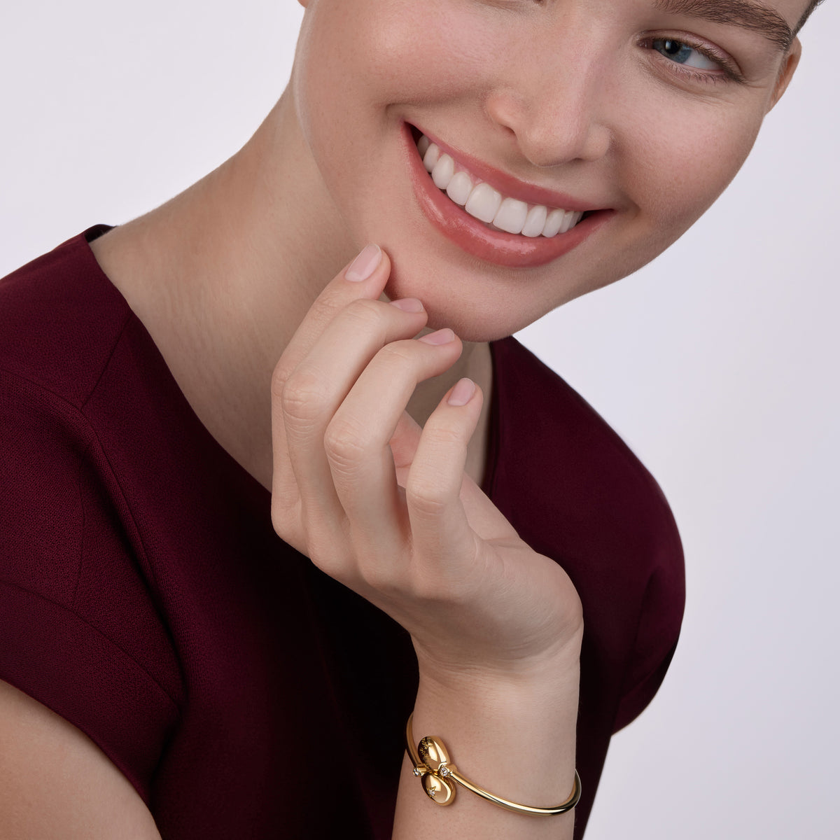 Close-up of a smiling model wearing the Heritage Gold Bangle Forever "I Love You" Crossover Bracelet in yellow gold, styled with a deep burgundy dress against a soft neutral background.