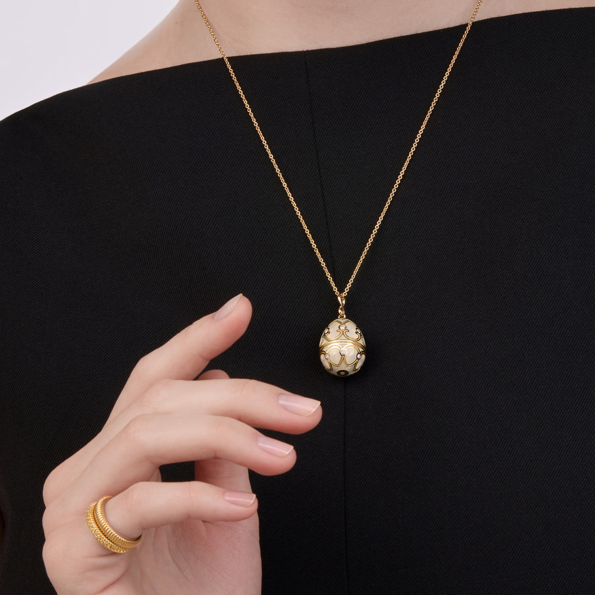 Close-up of Fabergé Heritage egg pendant necklace with white enamel and gold detailing, paired with a Love fluted gold ring on a model wearing a black dress.