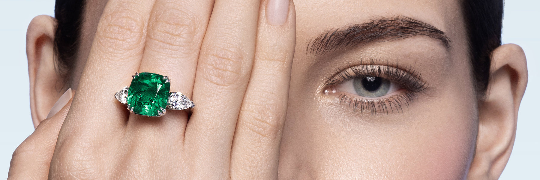 Close-up of a hand with a Fabergé emerald gemstone ring held near the face against a neutral background