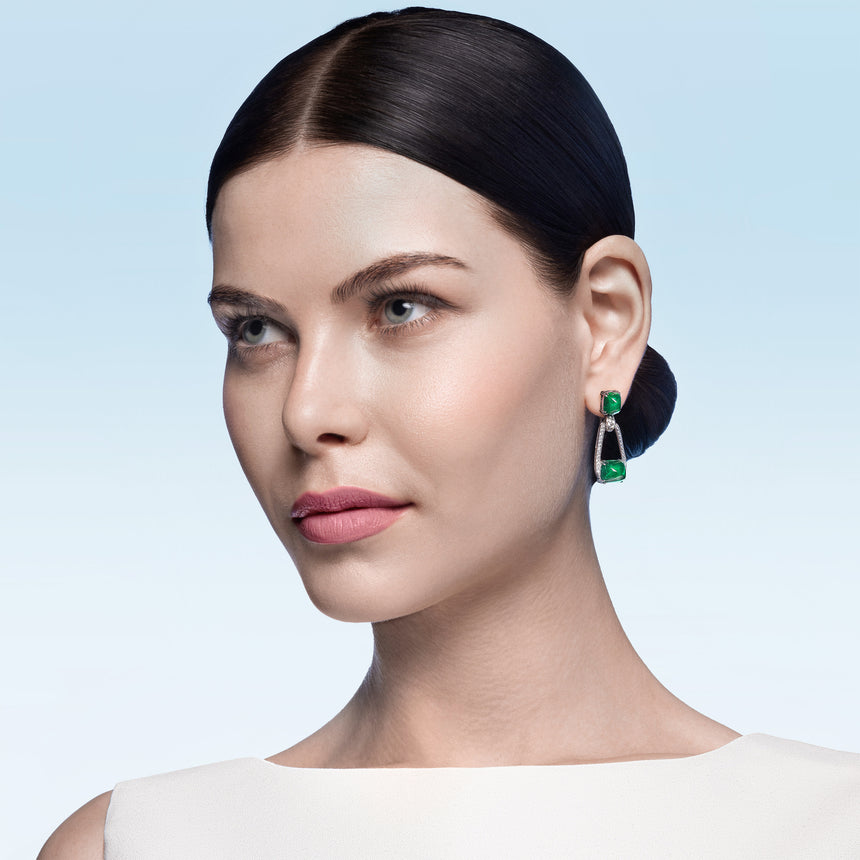High Jewellery category - Woman wearing green emerald and white gold earrings against a light blue background
