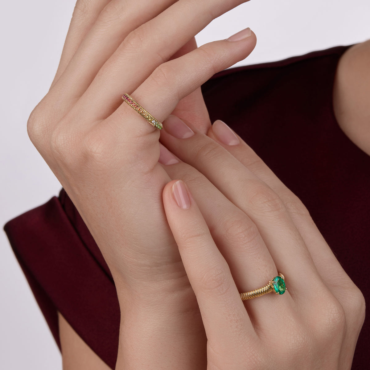 Model wearing two yellow gold Fabergé rings: an emerald pavé ring with diamonds and an eternity ring with mixed gemstones, styled against a burgundy satin sleeve.