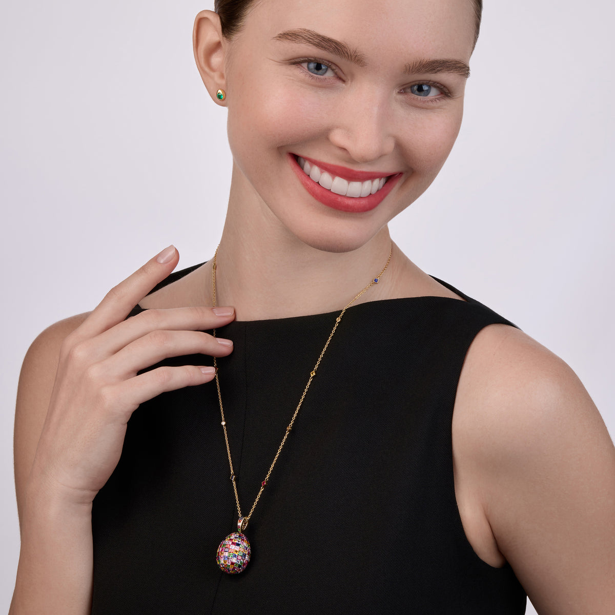 Model wearing the Fabergé Heritage Forever large egg pendant necklace with mixed gemstones, paired with Code emerald stud earrings in yellow gold.