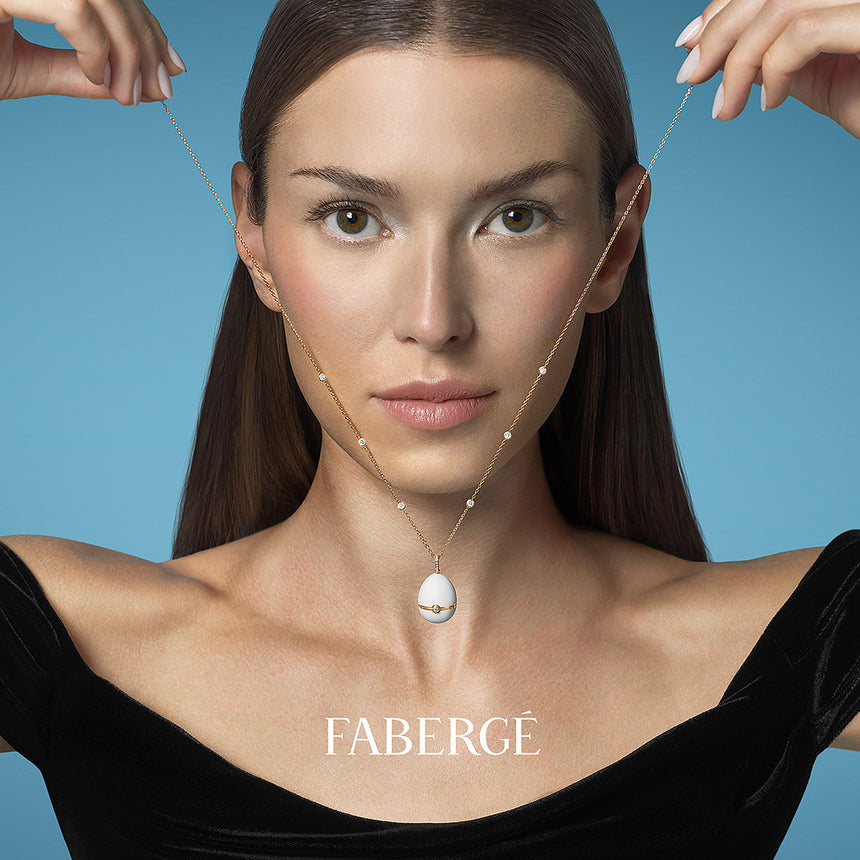 Model wearing the Fabergé №1 Egg Pendant in white enamel and yellow gold, holding the delicate chain on either side against a blue background.