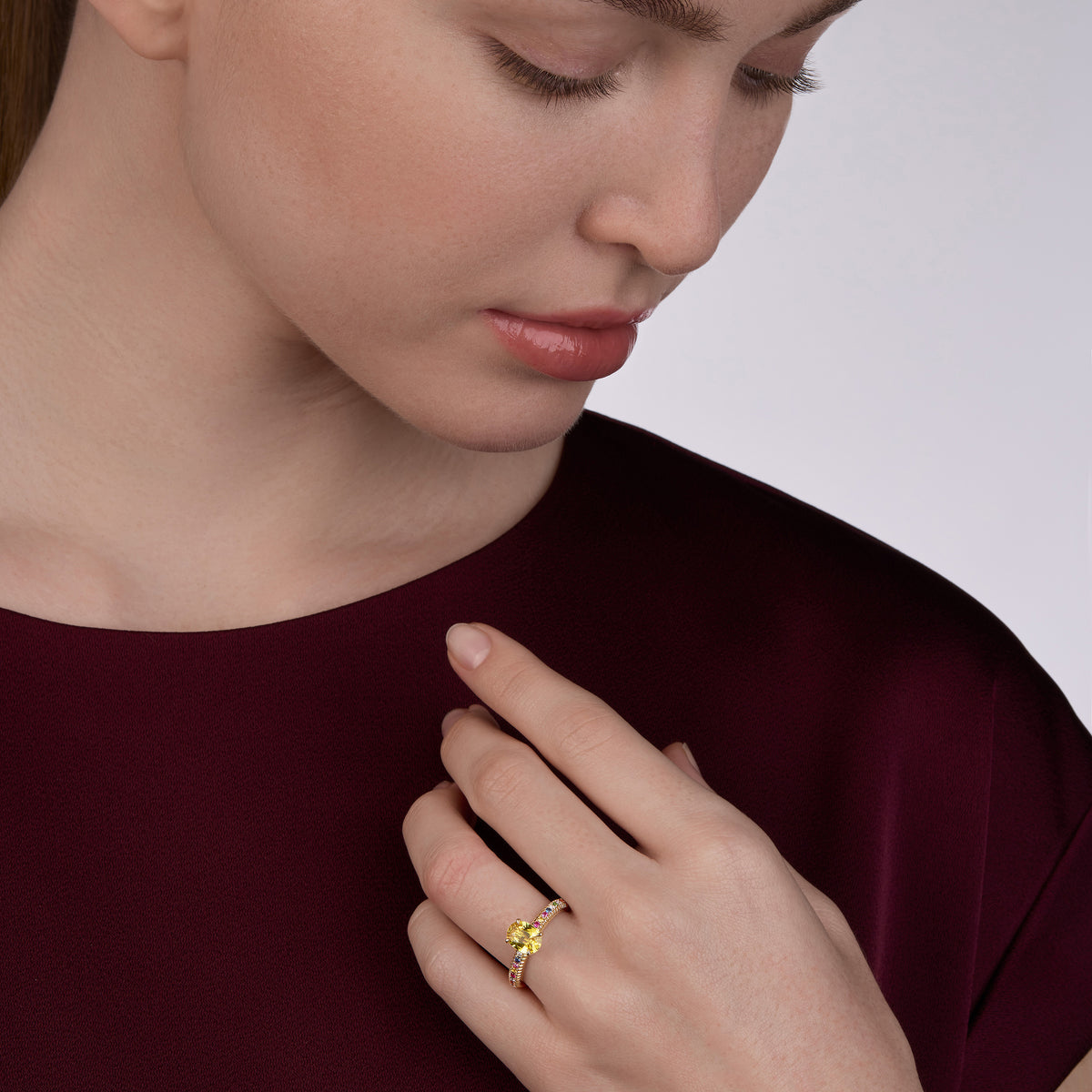 Close-up of a model wearing a yellow gold pavé ring featuring a yellow sapphire centre stone and mixed gemstones, styled against a deep burgundy dress.
